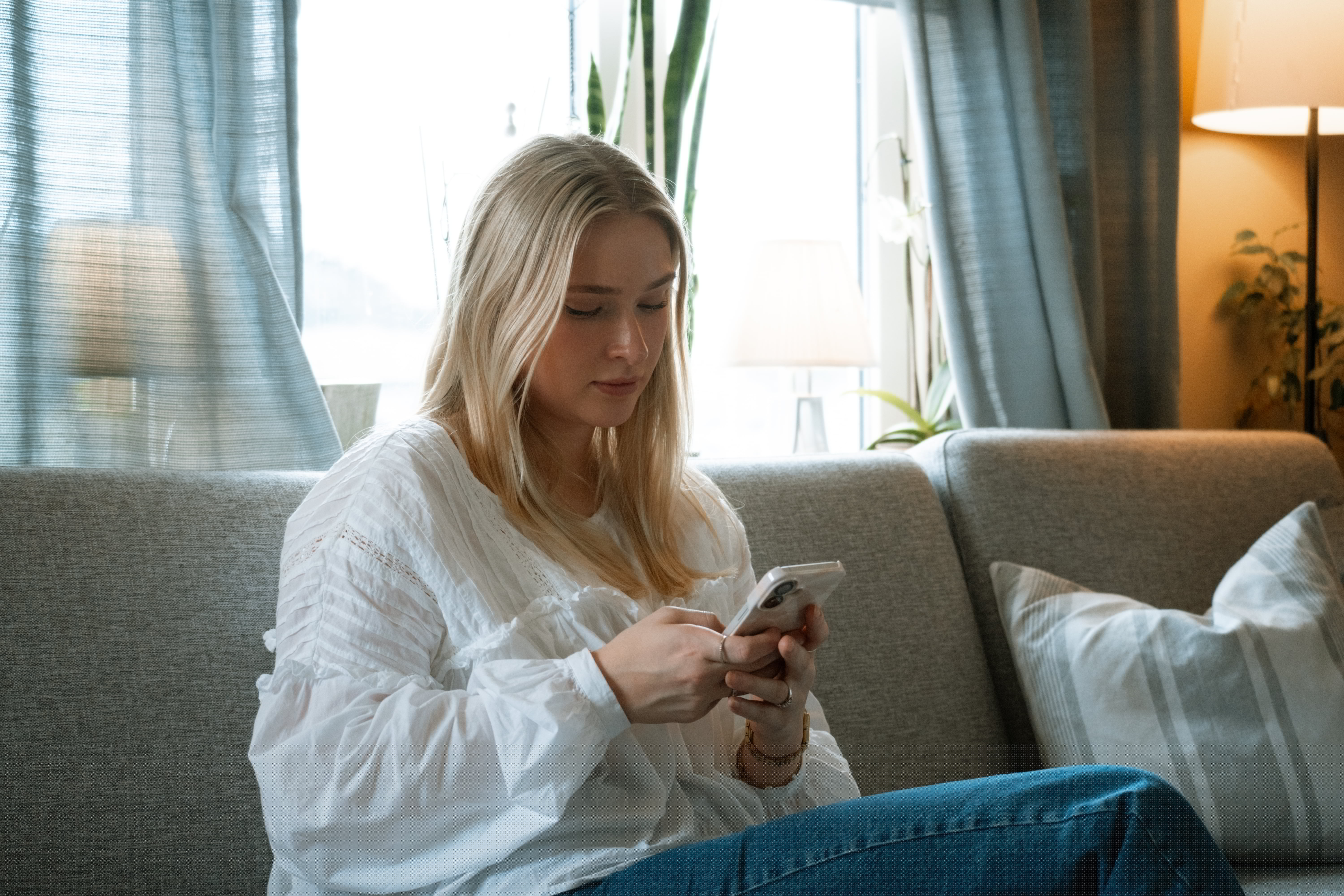 Person med hvit bluse sitter i sofaen og holder en mobil, med klokke på håndleddet.